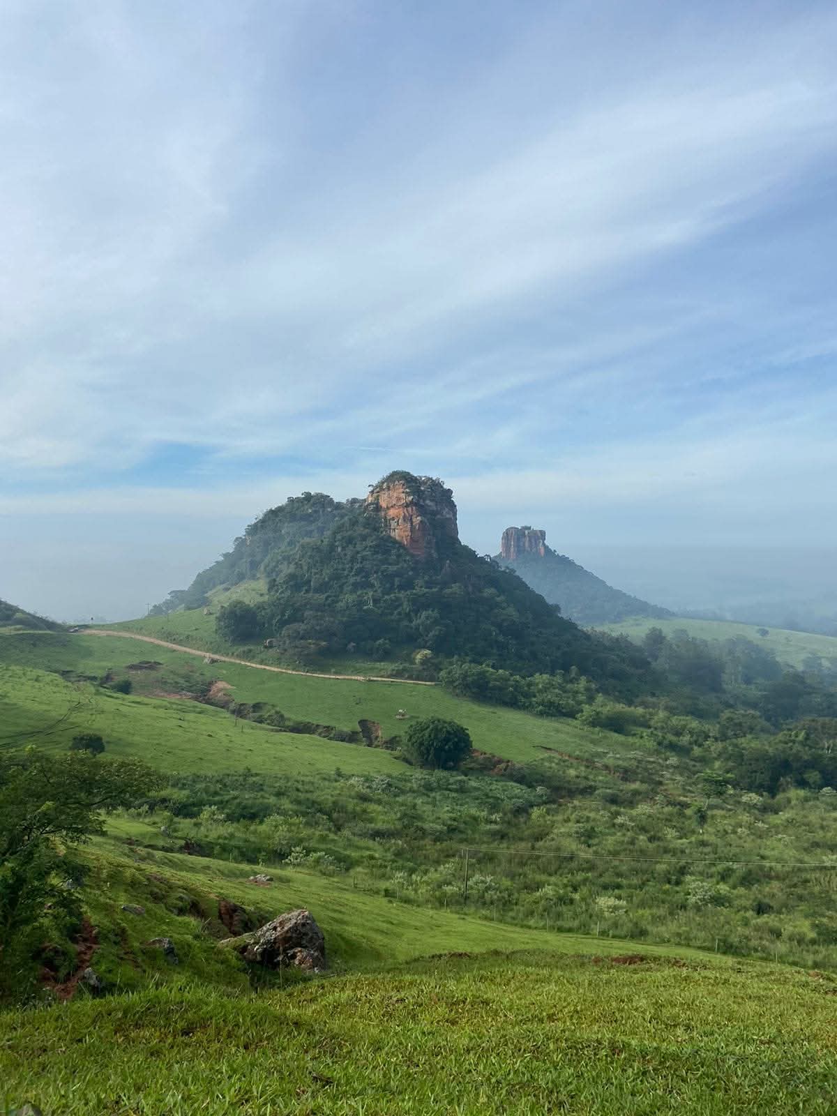 Morros do Camelo e do Cuscuzeiro vistos de dia, com estrada de terra, pastagens e névoa ao fundo