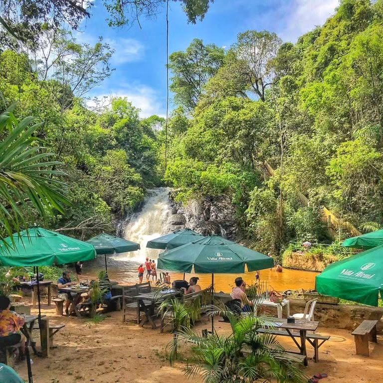Mesas e guarda-sóis do Cachoeira do Salto Bar e Restaurante montados na areia ao lado da Cachoeira do Salto Major Levy, em Analândia (SP), com a escadaria de corrimão amarelo na encosta à direita